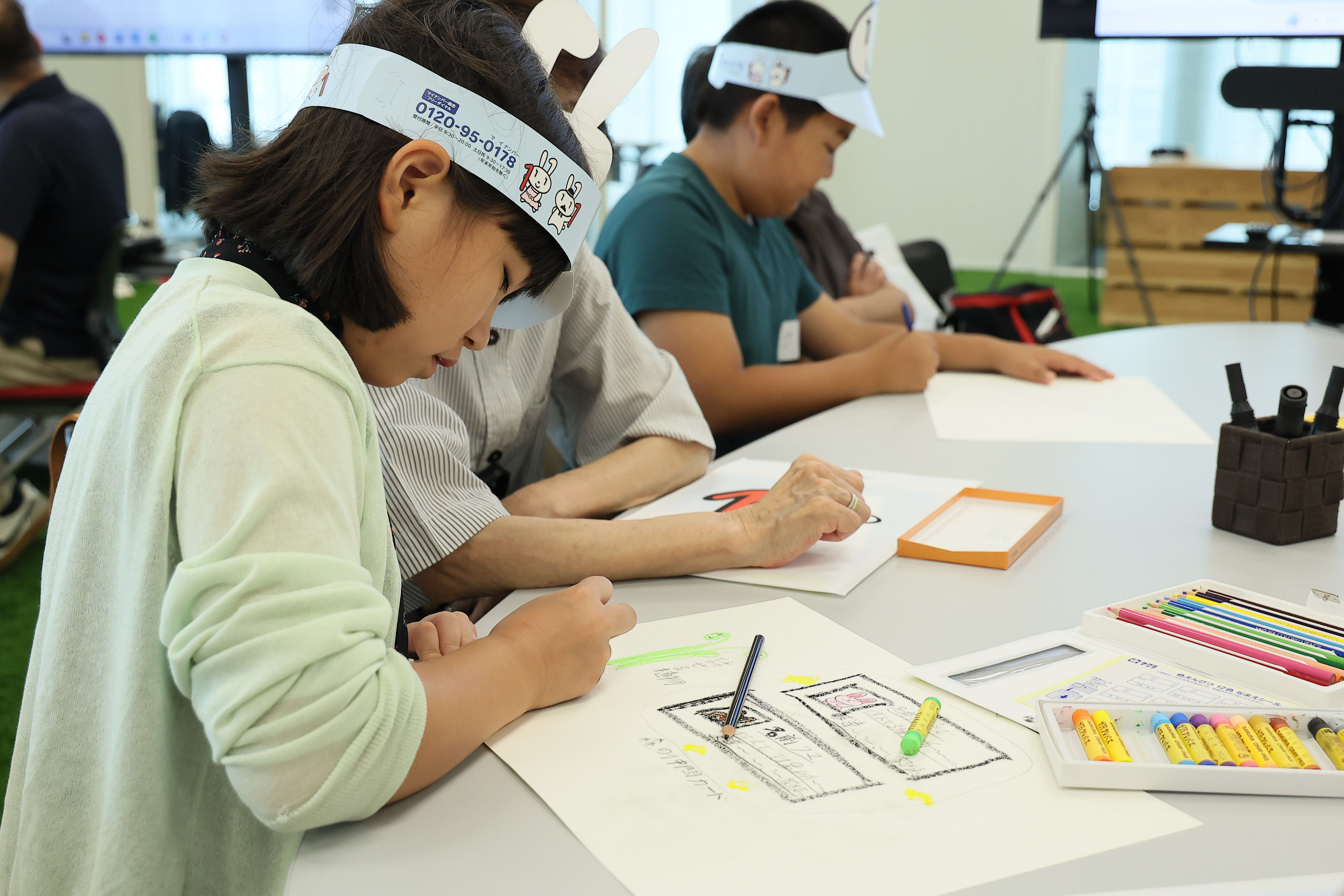 ワークショップ参加者の小学生3名が、クレヨンと色鉛筆を使って、生成AIを使って考えた「マイナちゃんの大冒険」の一場面を画用紙に描いている。小学生はマイナちゃんの紙製サンバイザーをかぶっている。