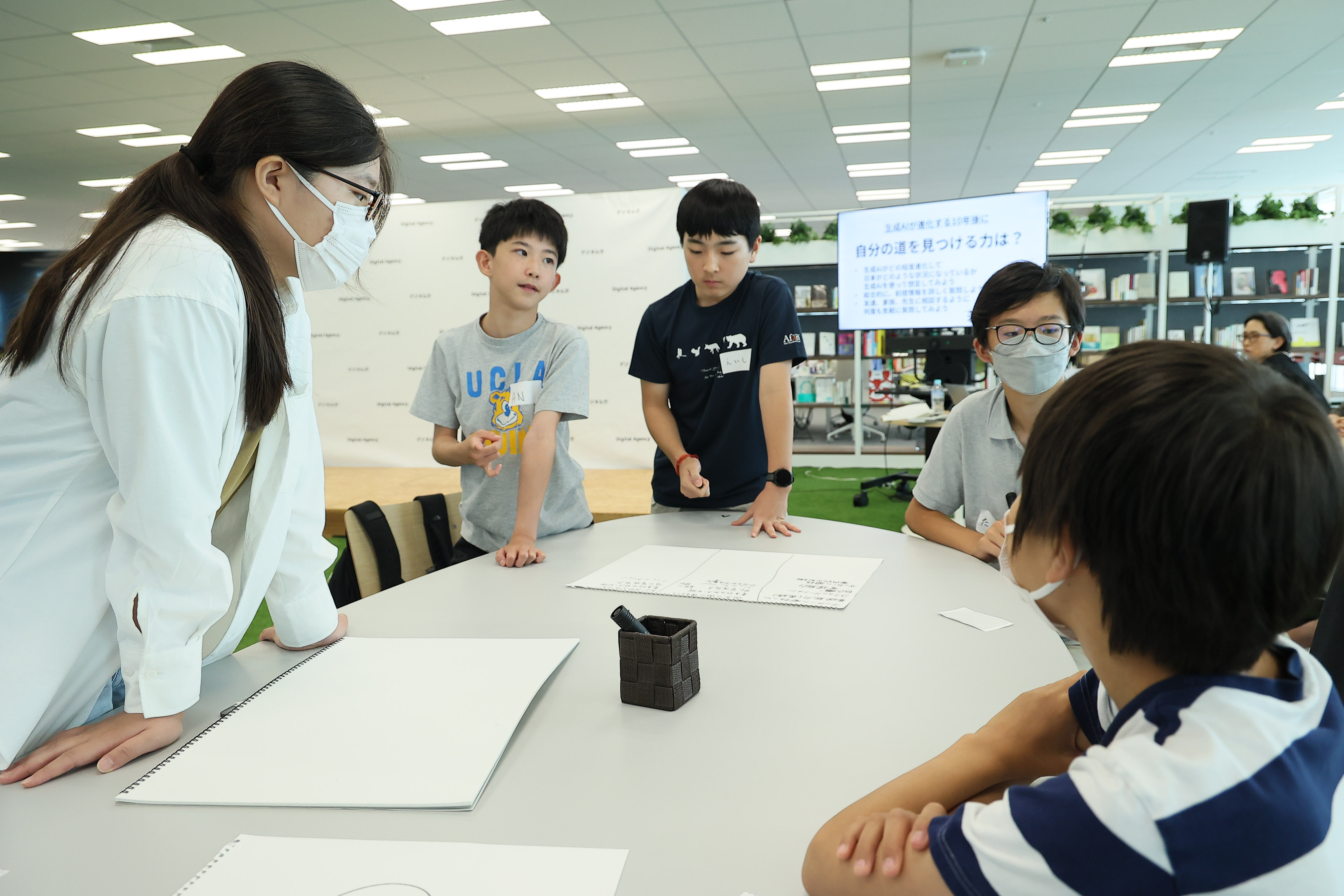 ワークショップ参加者の中学生5名で机を囲んで議論している。机の上にスケッチブック3つと黒色のペン1本が置いてある。背後のモニターには、議論のテーマが映し出されており、「 自分の道を見つける力は?」と記載されている。