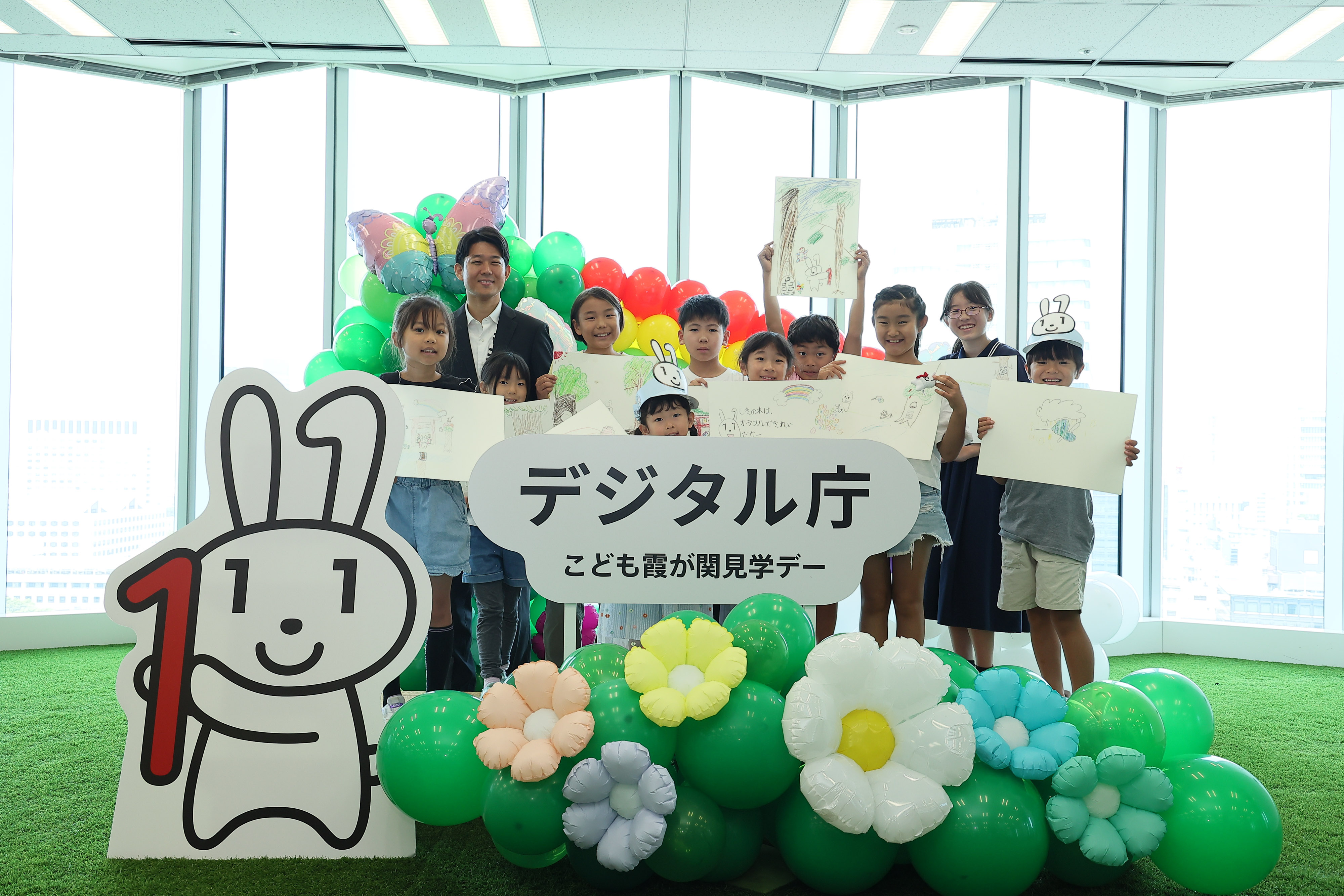 「デジタル庁 子ども霞が関見学デー」のロゴパネル、マイナちゃんのパネル、花や蝶のバルーンが飾られているフォトスポットに、ワークショップに参加した小学生と岸デジタル大臣政務官が立っている。小学生は、生成AIをつかって考えた「マイナちゃんの大冒険」の一場面を描いた画用紙を掲げている。