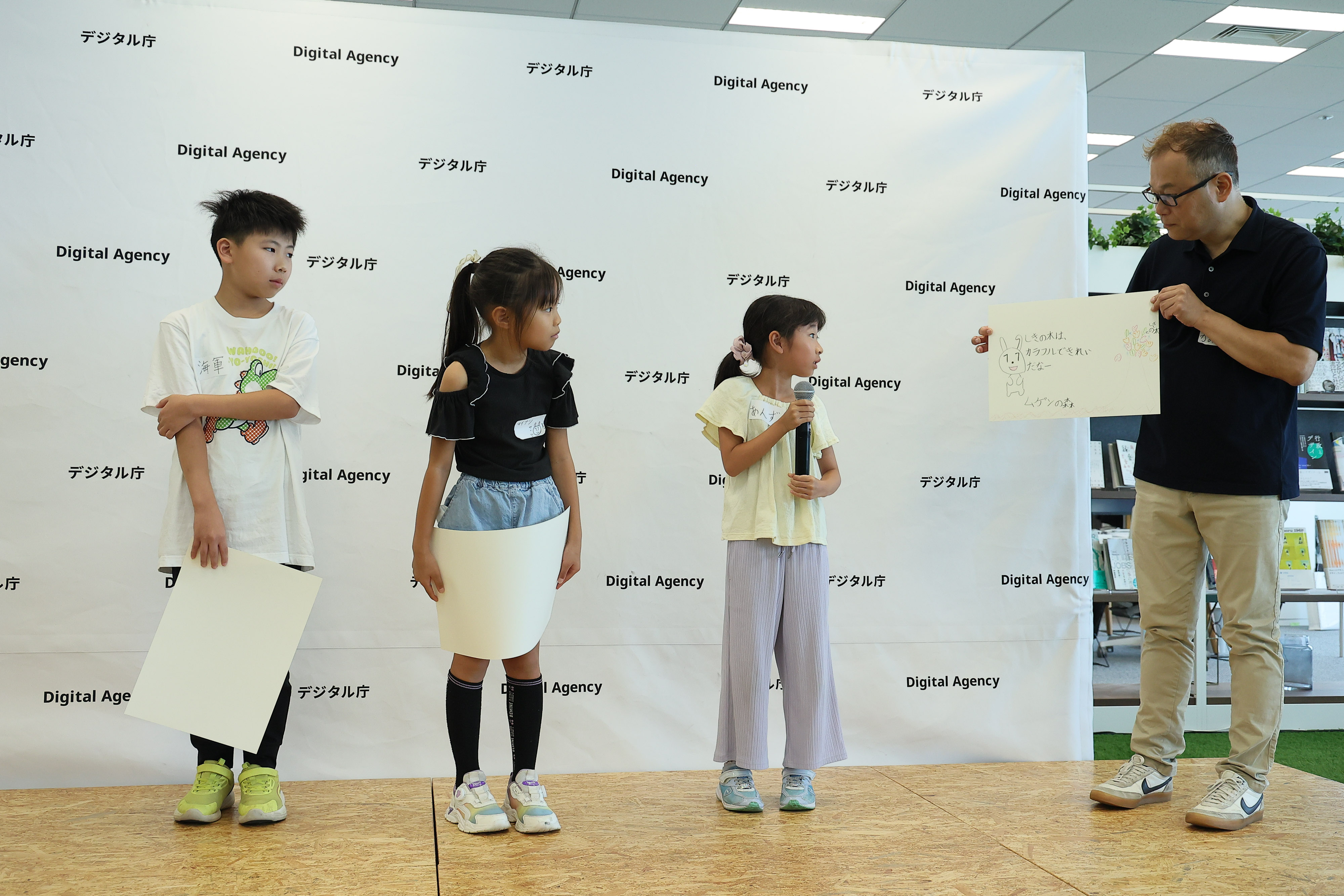 ワークショップ参加者の小学生3名が、デジタル庁のロゴをあしらったパネルが設置されたステージにたっている。デジタル庁職員が右側に立ち、小学生のうち1名が描いた「マイナちゃんの大冒険」物語の絵を手に持っている。その小学生は、マイクを持ち絵の説明をしている。小学生2名は、描いた絵を手に持ち発表されている絵を見ている。