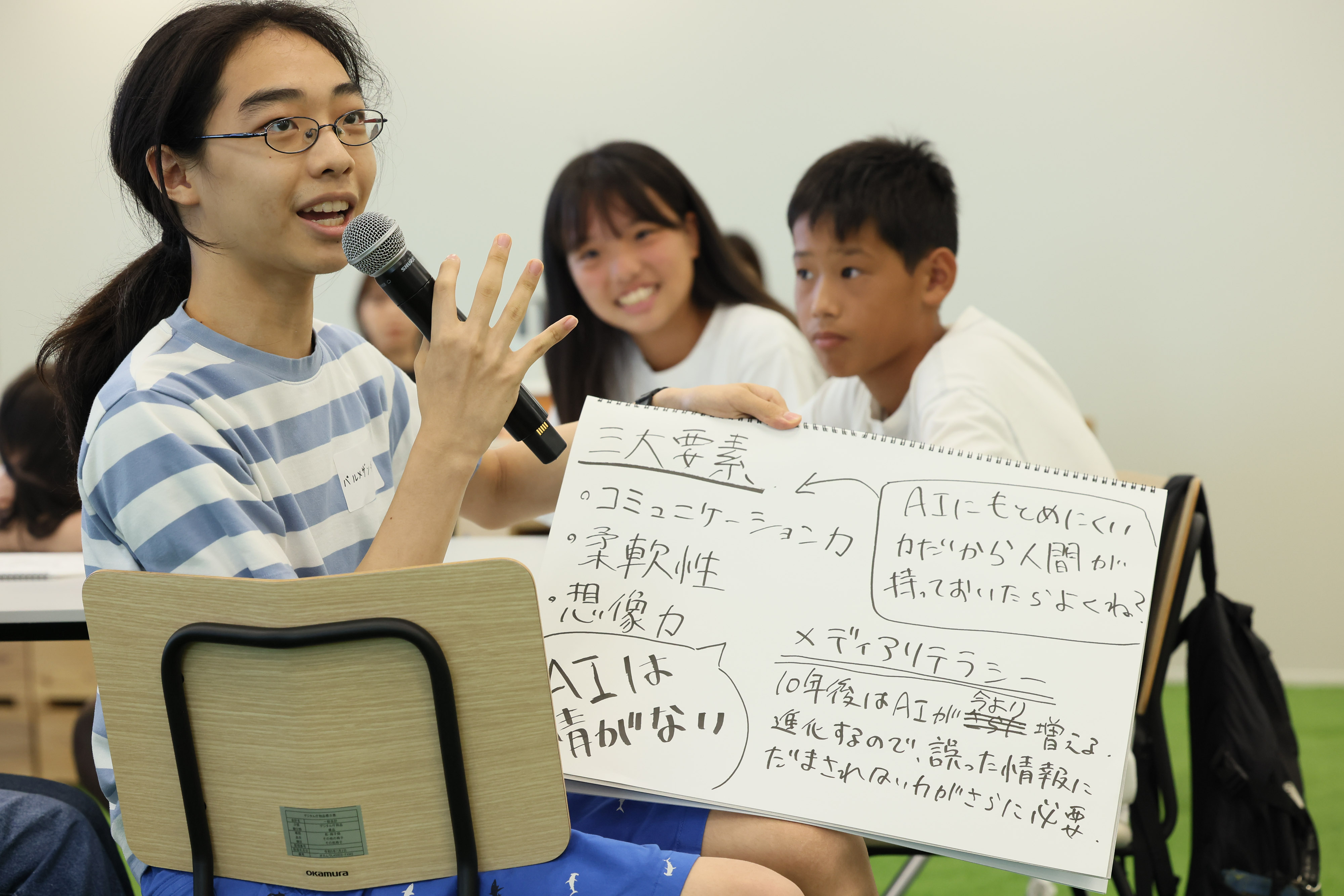 ワークショップ参加者の中学生3名のうち、1名が議論の内容をメモにまとめたスケッチブックを手に持ちマイクで発表している。中学生2名は、スケッチブックを手に持って発表している中学生を見ている。