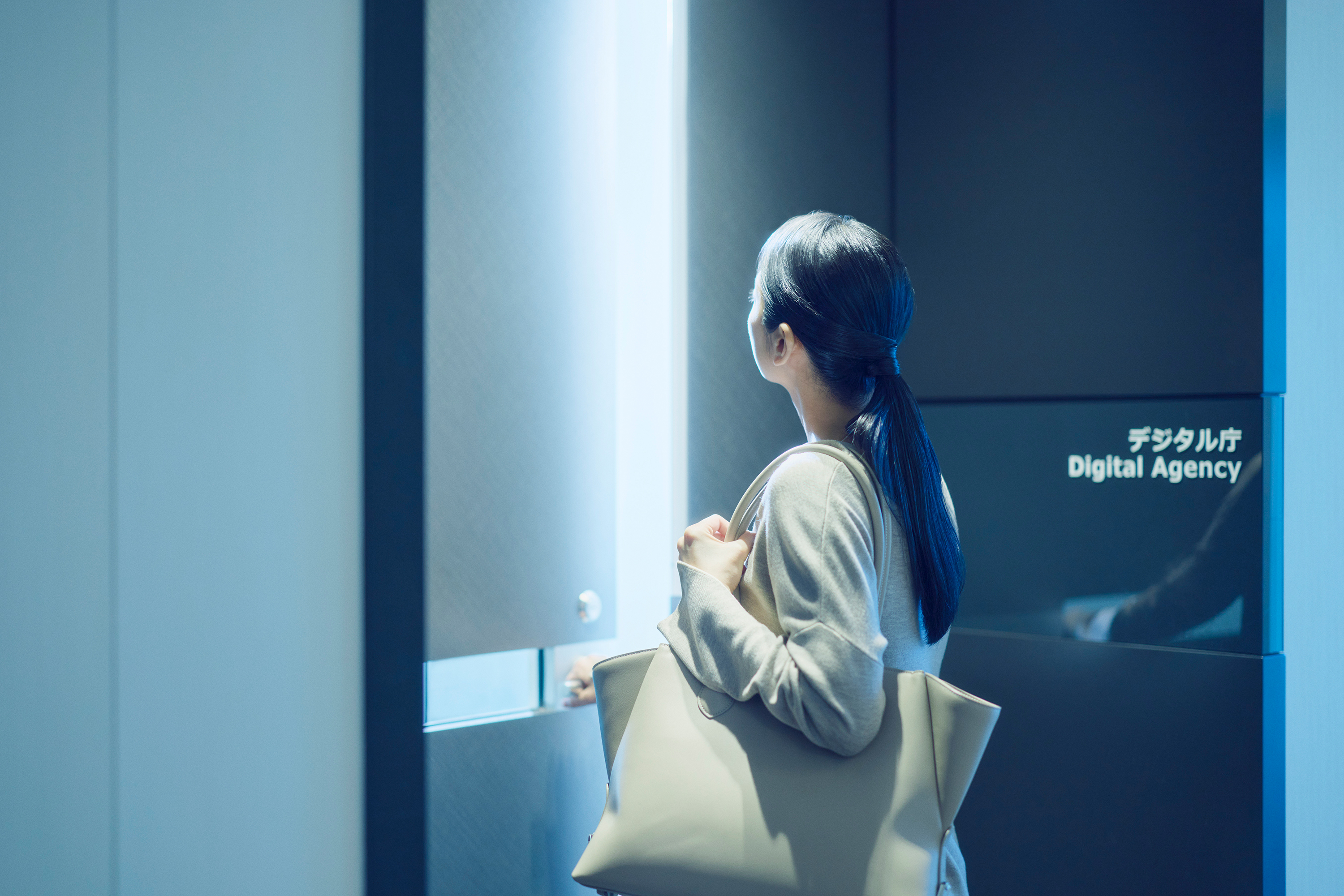 A photograph of the rear view of a woman standing in front of the entrance to the Digital Agency office. The woman is facing the entrance and is about to enter, with her right hand on the doorknob. She has long black hair pulled back into a ponytail, is wearing a beige knit, and has a white tote bag slung over her shoulder. On the front right, you can see a black wall with the words "Digital Agency Digital Agency" written in both Japanese and English. Light is shining from inside the office.