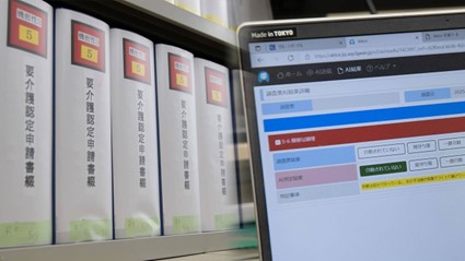 2枚の写真を組み合わせて構成。左側の写真では「要介護認定申請書綴」という背表紙タイトルが付いた、書類ファイルが5冊収められた書類棚を写している。右側の写真では、ノートパソコンの画面が写されており、要介護認定事務AIシステムの画面が投影されている。