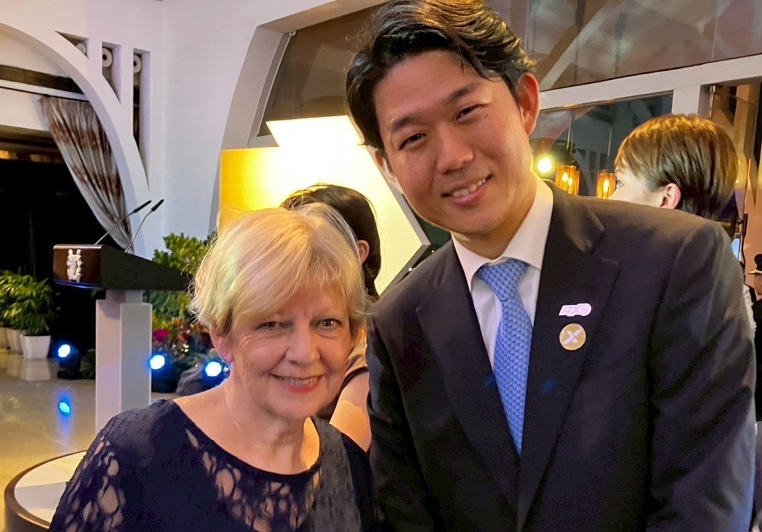 二人が並んで立っている写真。左側の人物は紺色の服を着用している。右側の人物は紺のスーツと水色のネクタイを着用し、胸にピンバッジを付けている。両者とも笑顔で、背景には植物や照明が見える。