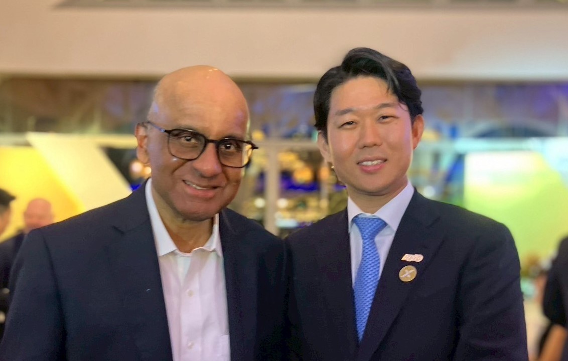 二人が並んで立っている写真。左側の人物は眼鏡をかけ、紺のジャケットとピンクのシャツを着用。右側の人物は紺のスーツと水色のネクタイを着用し、胸にピンバッジを付けている。