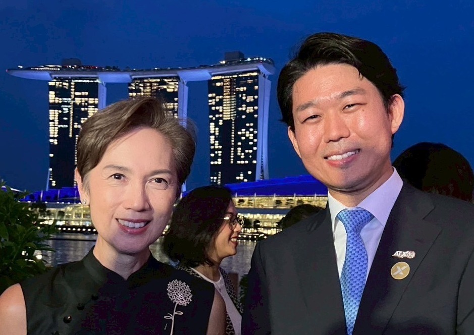 二人が並んで立っている写真。左側の人物は黒い服を着て、胸元に花の刺繍がある。右側の人物は紺のスーツと水色のネクタイを着用し、胸にピンバッジを付けている。両者とも笑顔で、夜景を背景に建物が映っている。