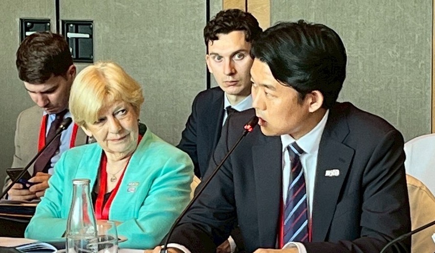 セッション中の写真。テーブルに着席した複数の参加者が映っている。左側には緑色のジャケットを着た人物が座っており、その右側で濃紺のスーツを着た岸デジタル大臣政務官が発言している。テーブルの前には参加者の名札が置かれている。