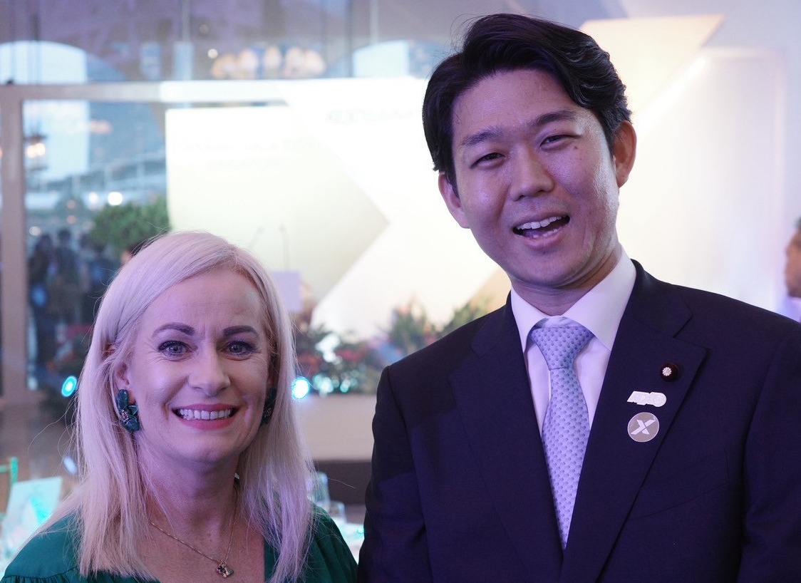 笑顔で並んで立つ二人の写真。左側の人物は緑色の服を着ており、右側の人物はスーツを着ている。背景には植物の装飾が見える。