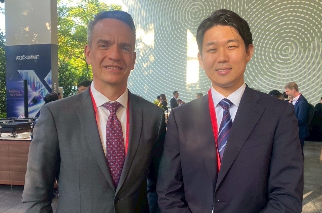 二人が並んで立っている写真。左側の人物はグレーのスーツと赤系のネクタイを着用している。右側の人物、紺のスーツと青いネクタイを着用している。両者とも赤い首掛けのIDカードを付けており、笑顔で立っている。背景には木々や建物の一部が見える。