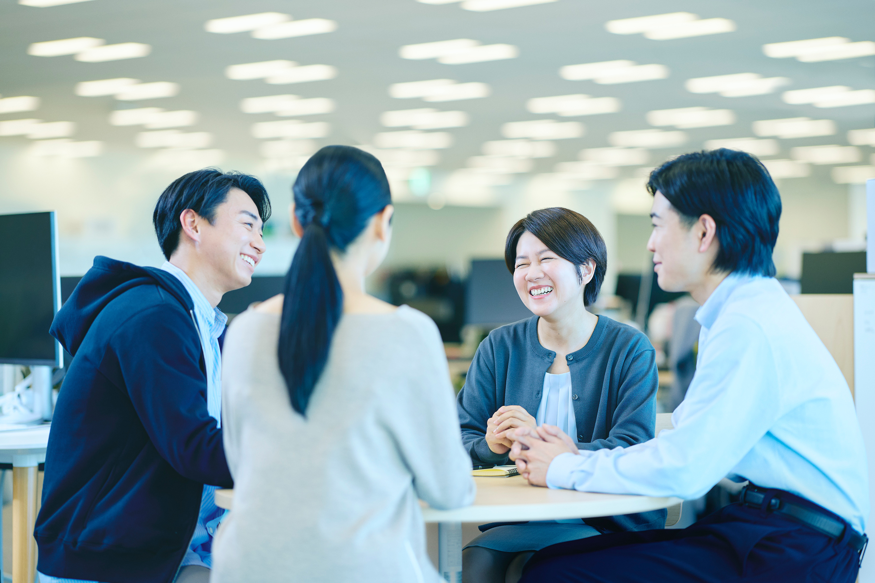 明るいオフィスでテーブルを囲み議論する男性2名・女性2名の計4名。1名は後ろ姿で、他の3名は互いに積極的に関わり合っており、共同ミーティングまたはブレインストーミングの様子。