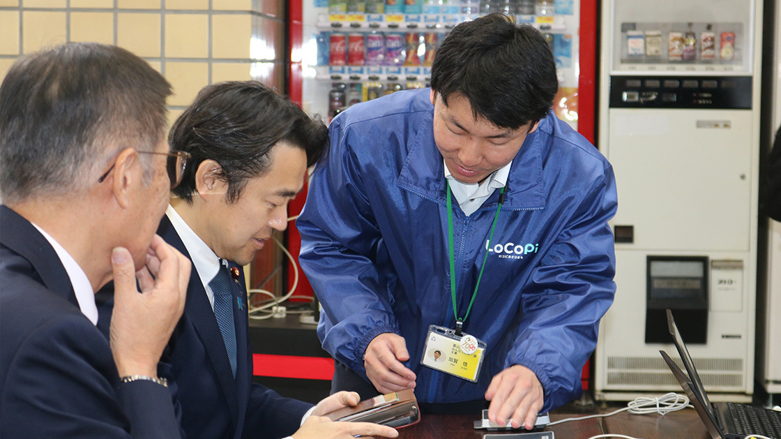 Mr. Hosaka, State Minister for Digital Transformation, is registering to use LoCoPi Asahimachi using a smartphone while receiving an explanation from a Asahimachi official. 