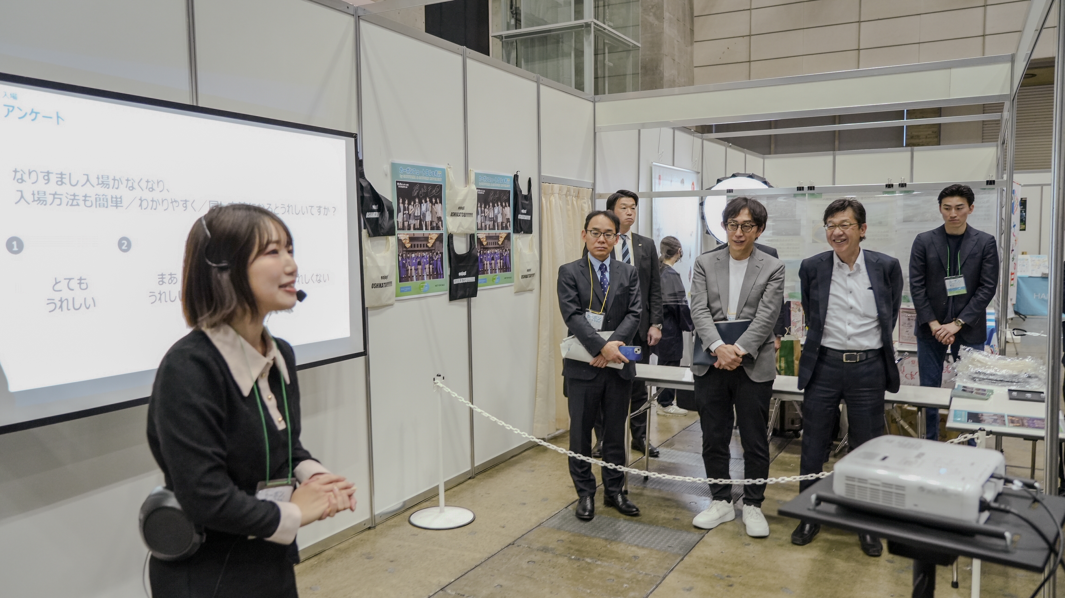 会場のブース内で、黒い服にヘッドセットマイクを着けた若い女性スタッフが説明を行っている。背後のスクリーンには「アンケート」と題したスライドが映し出されており、「なりすまし入場がなくなり、入場方法も簡単/わかりやすく/同じ…とうれしいですか?」という設問と、「①とてもうれしい」「②まあうれしい」などの選択肢が表示されている。ロープの内側にはスーツ姿の男性3名が並んで聴講している。ブース内にはグッズや写真パネルが展示されている。
