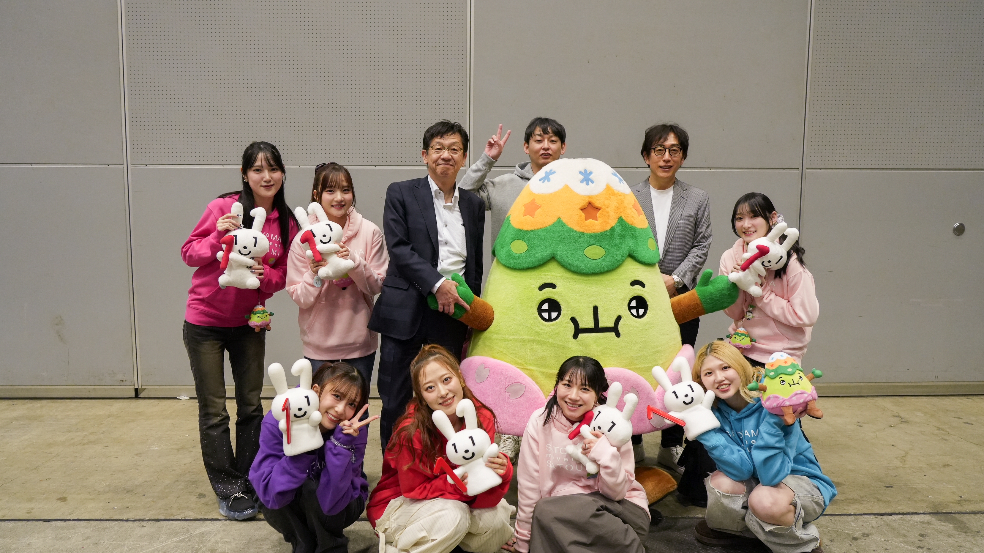 屋内施設の灰色の壁を背景に、計10名が集合写真を撮影している。中央には黄緑色の山型をした大きな着ぐるみのキャラクター「さとやまくん」が立ち、その周囲をピンク・紫・水色などのカラフルなパーカーを着た女性たちが囲んでいる。後列中央にはスーツ姿の男性2名が立っており、全員が笑顔でポーズをとっている。女性たちの多くは白いマイナちゃんのぬいぐるみを手に持っている。