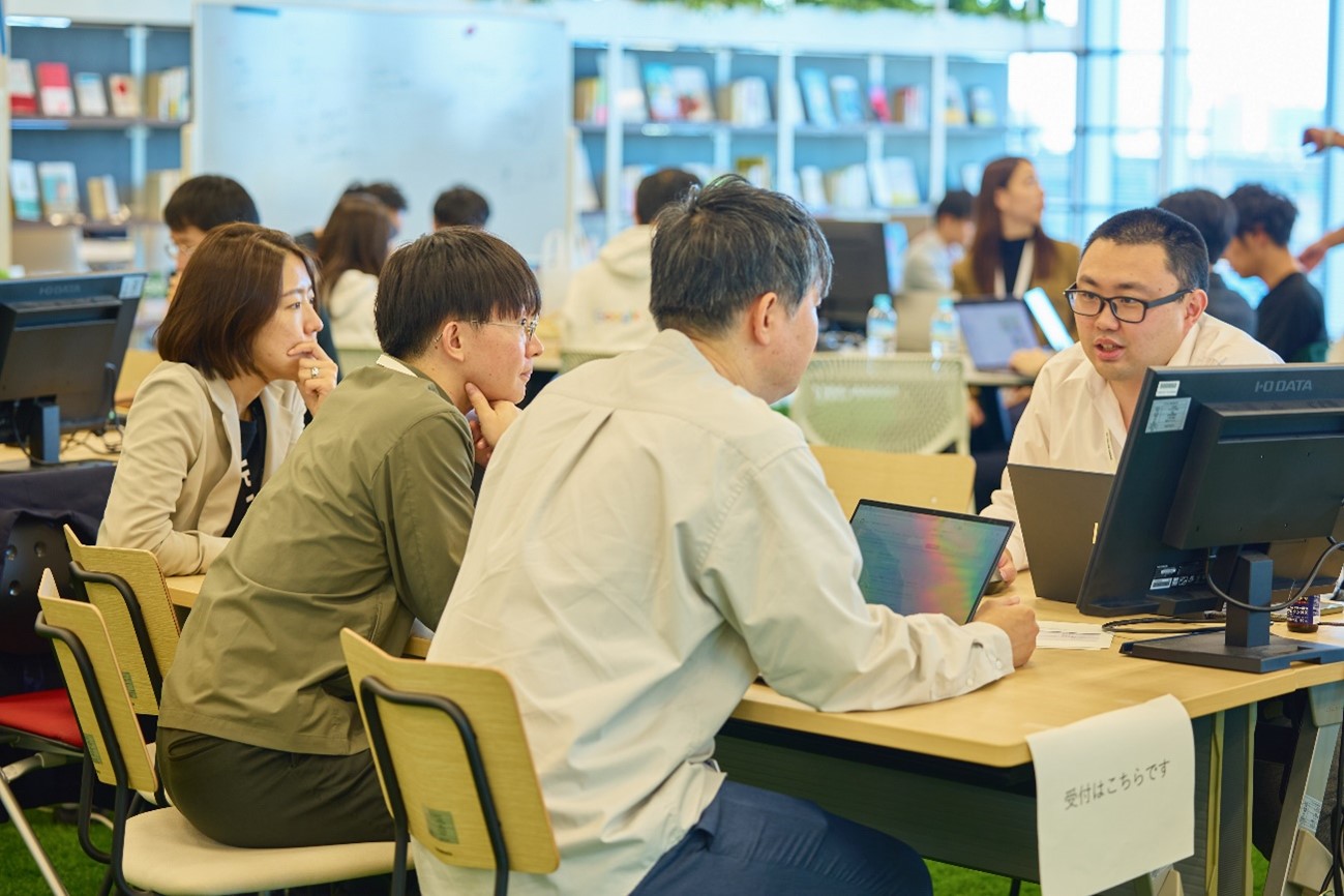 The three government officials are seated side by side around the table, and are talking with the person in charge of the consultation counter sitting across from them. It seems that government officials is talking about the development of a prototype of AI as a business improvement, bringing in his daily worries. 