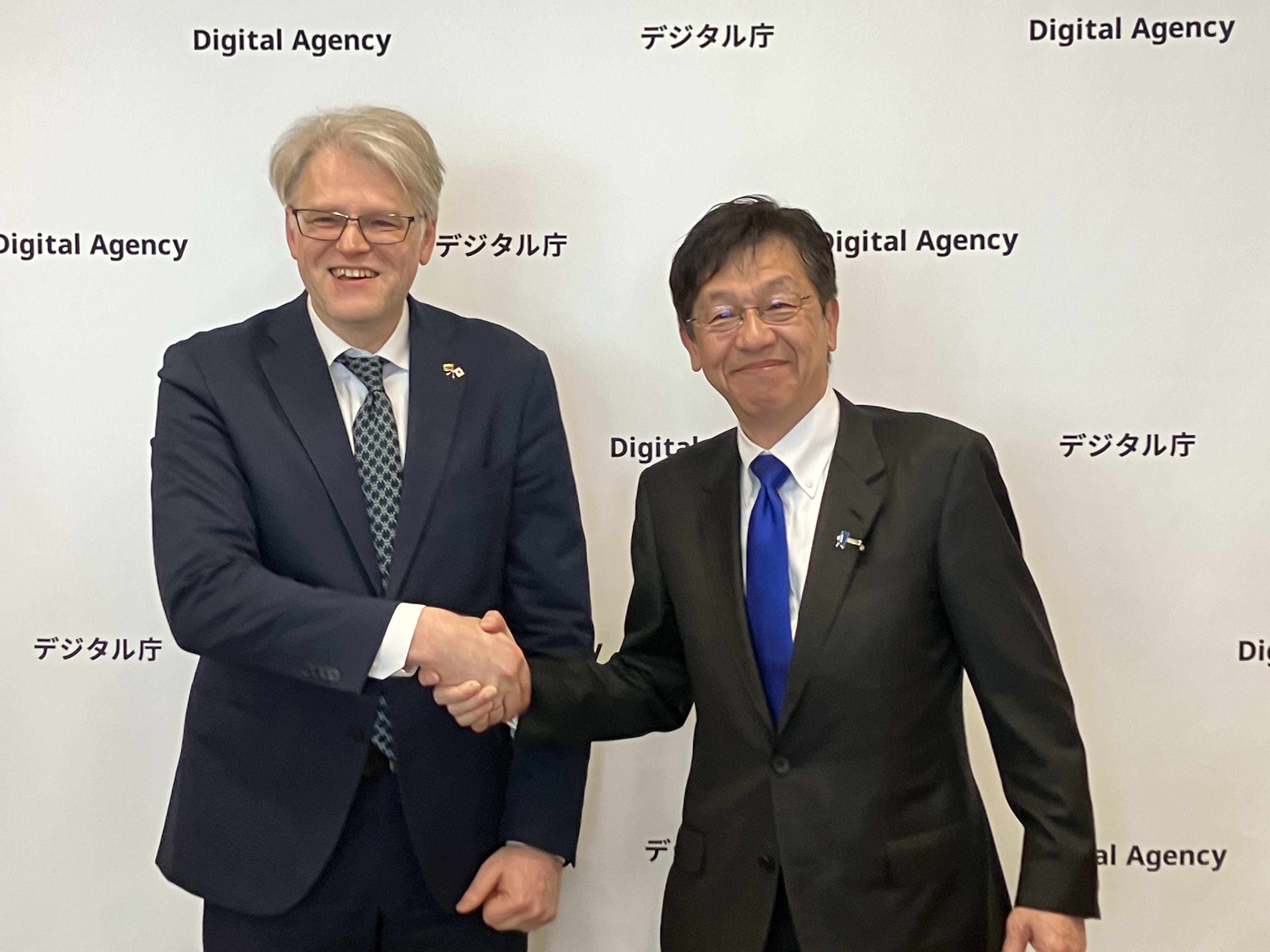 リトアニア共和国駐日大使のオーレリウス・ジーカス氏(左)と、松本デジタル大臣(右)が、デジタル庁のロゴが繰り返し表示されたバックパネルの前で握手している様子