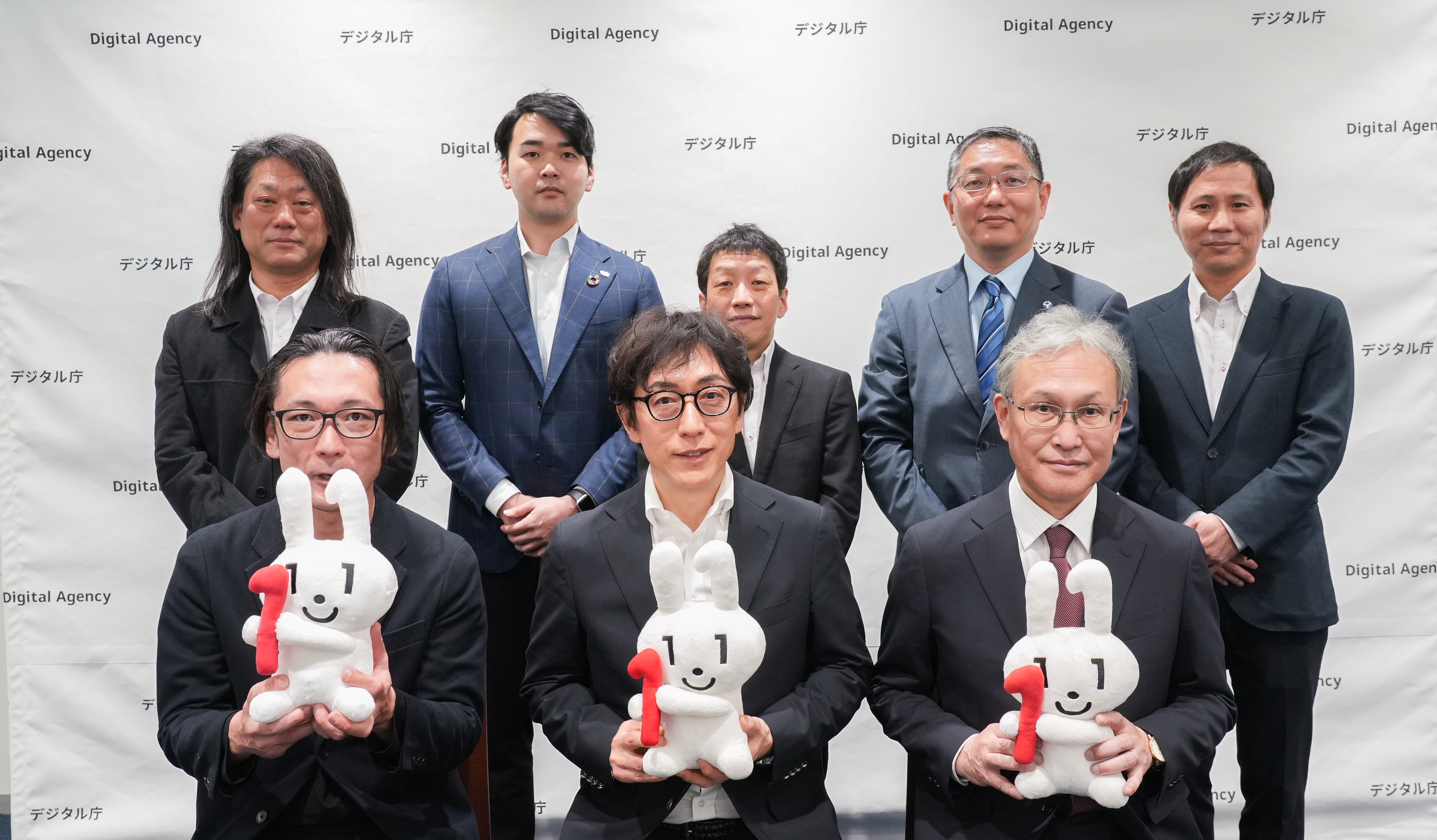 デジタル庁のロゴが繰り返し配置された白い背景の前で、スーツ姿の男性8名が2列に並んだ集合写真。後列左から一般社団法人OpenIDファウンデーションジャパン 富士榮尚寛代表理事、株式会社コシダカホールディングス 腰髙理志執行役員、大和大学 田中秀幸教授、国立大学法人大阪大学 鎗水徹教授、国立大学法人広島大学 渡邉英伸准教授。前列左から西日本旅客鉄道株式会社 眞鍋宗一郎課長、デジタル庁国民向けサービスグループ 鳥山高典企画調整官、国立情報学研究所 佐藤周行教授が立っている。前列の3名はデジタル庁のマスコットキャラクター、マイナちゃんが赤い「1」の数字を持ったぬいぐるみを手に持っている。