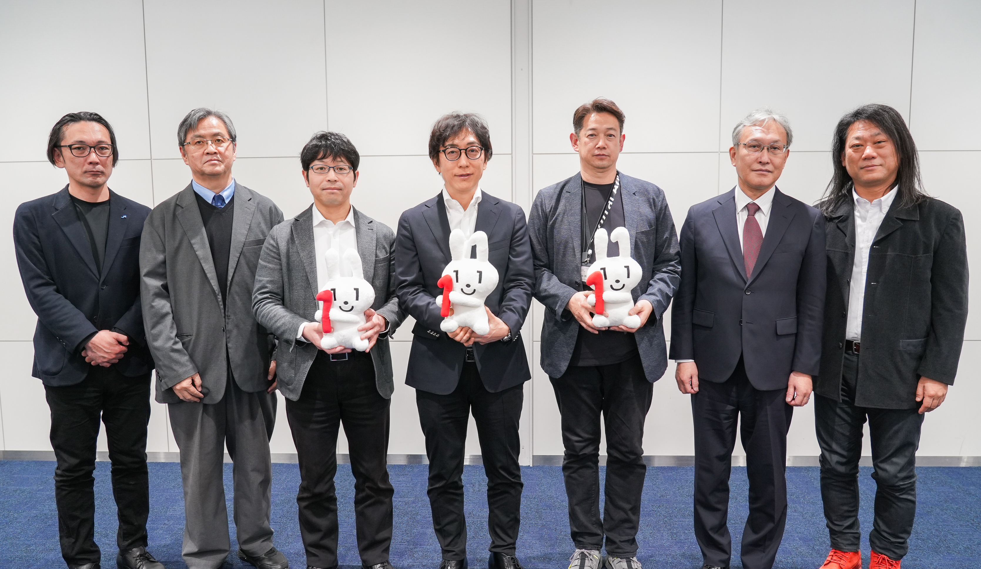 シンポジウムにて参加者が記念撮影する様子。7名の男性が横一列に並んで記念撮影している。左から西日本旅客鉄道株式会社 眞鍋宗一郎課長、順天堂大学 満塩尚文准教授、デジタル庁国民向けサービスグループ 久芳全晴企画官、デジタル庁国民向けサービスグループ 鳥山高典企画調整官、デジタル庁デジタル社会共通機能グループ 小畑雅人参事官補佐、国立情報学研究所 佐藤周行教授、一般社団法人OpenIDファウンデーションジャパン 富士榮尚寛代表理事が立っている。中央の3名が赤い「1」の数字が描かれたマイナちゃんのぬいぐるみを手に持っている。