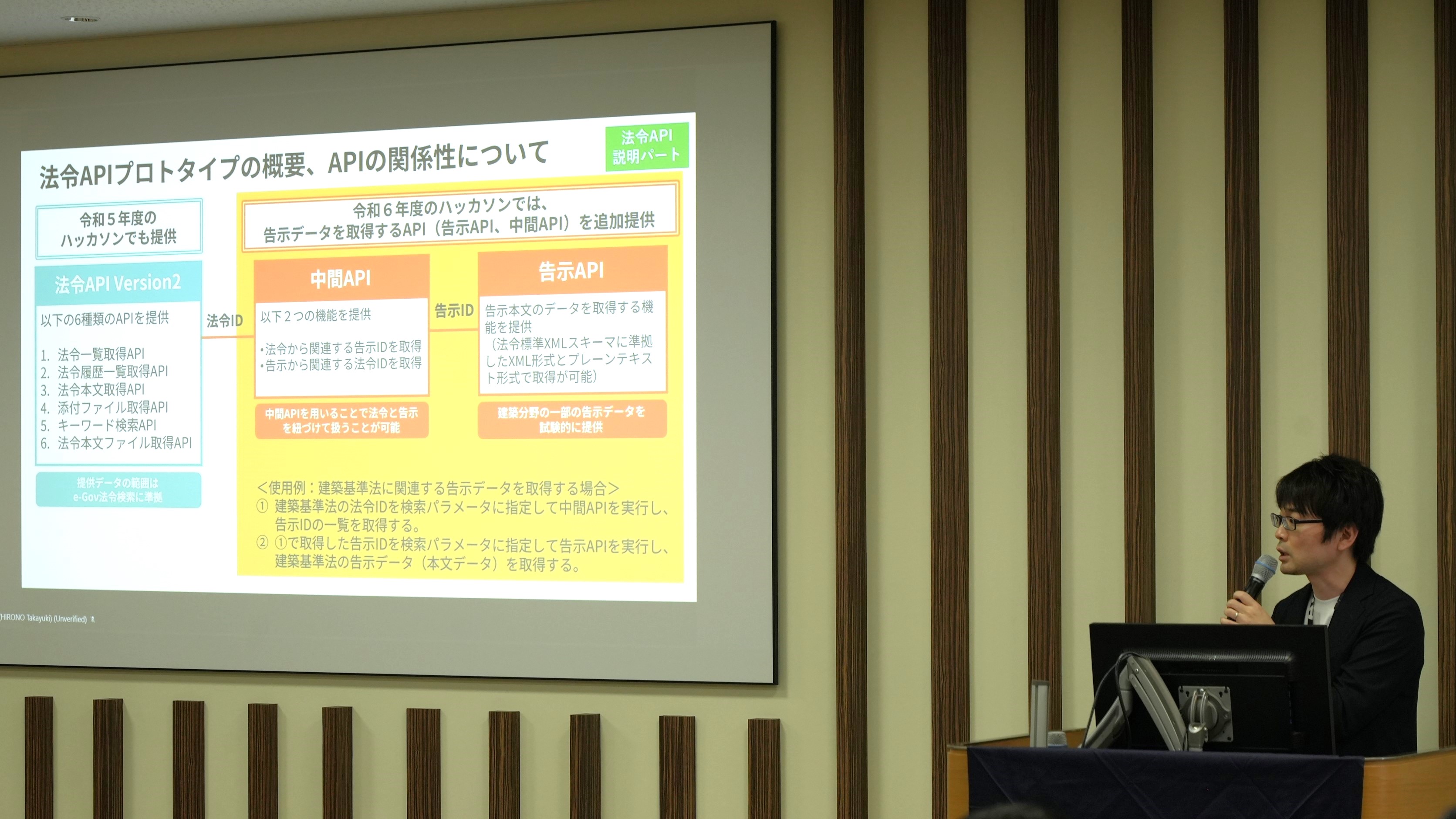 Hirono, the system engineer of the Digital Agency, explains the system of Digital Agency. On the screen beside Hirono, an outline of the laws and ordinances API-Protocol is projected. 