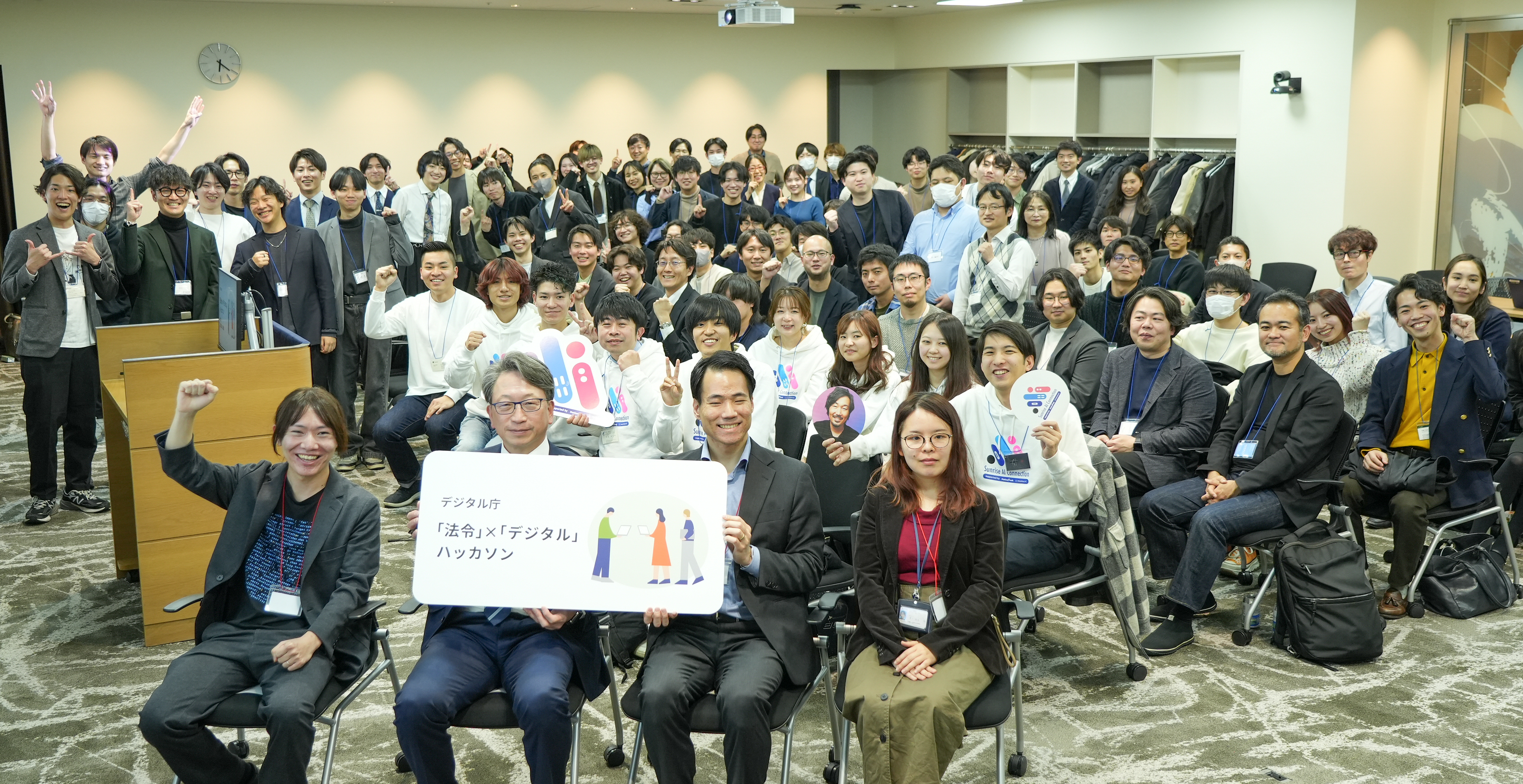 A group photo of Mr. Taira, Minister for Digital Transformation,, three judges, and the hackathon participants. Mr. Taira, Minister for Digital Transformation, and three judges are seated in the front row, with Mr. Taira, Minister for Digital Transformation, and Mr. Yagita, the judge, holding a board marked 