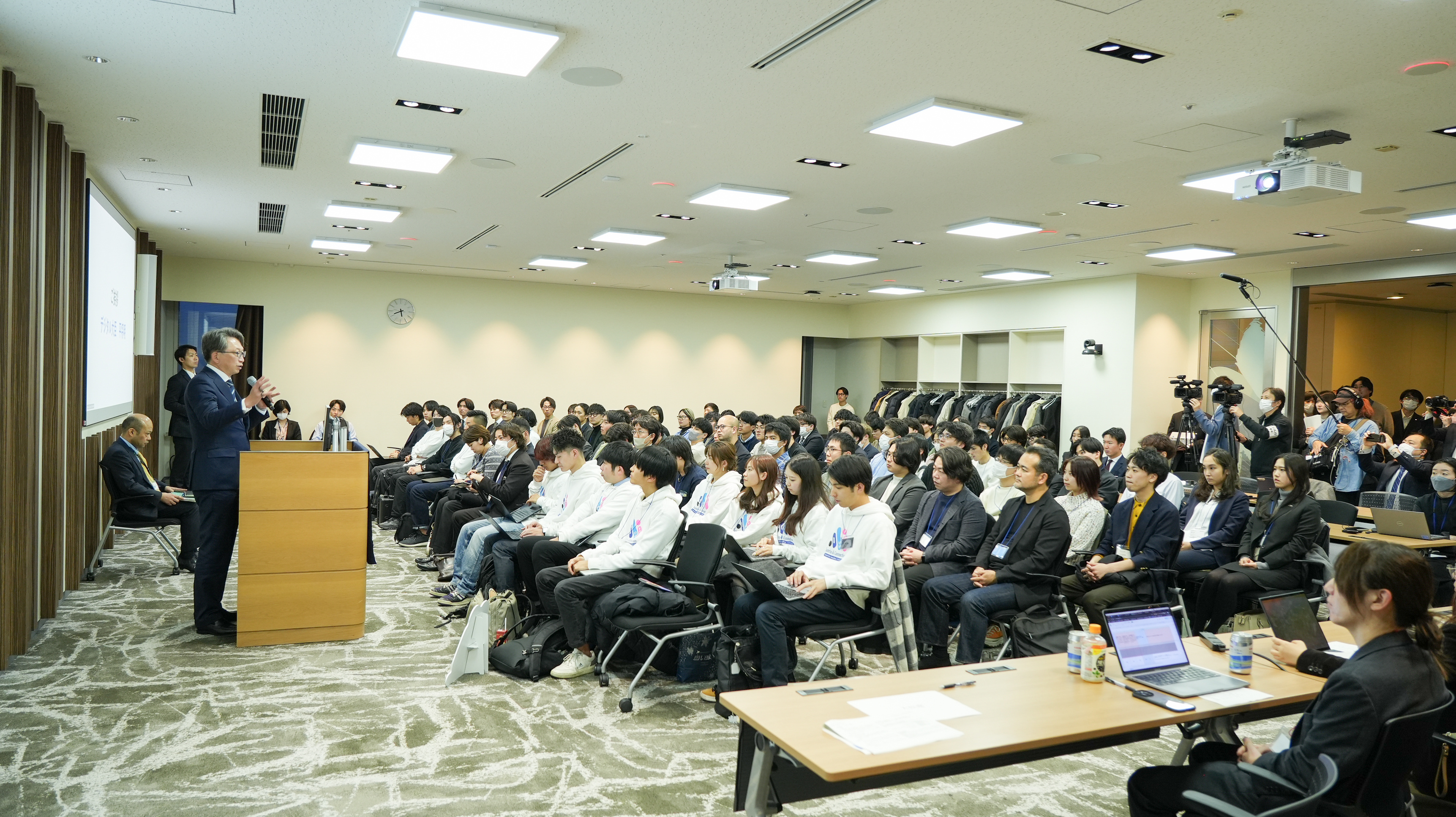 A scene of the entire Hackathon venue where Minister Taira delivered a speech from the platform. Approximately 100 participants were seated and listened to Minister Taira. 